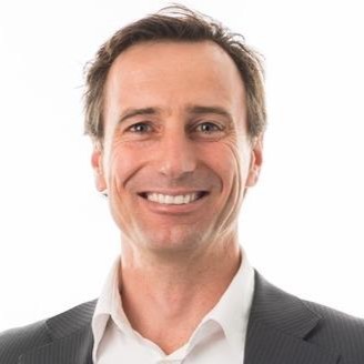 Smiling man with short brown hair wearing a white shirt and dark suit jacket against a white background.