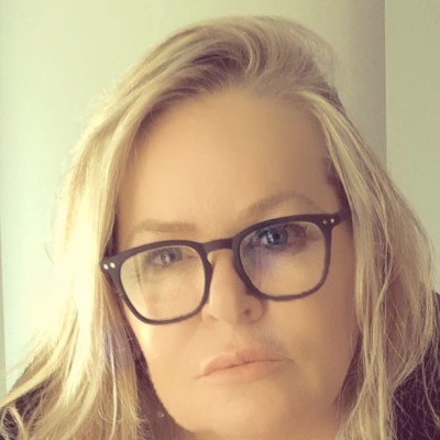 Close-up of a woman with blonde hair and black-framed glasses looking directly at the camera.